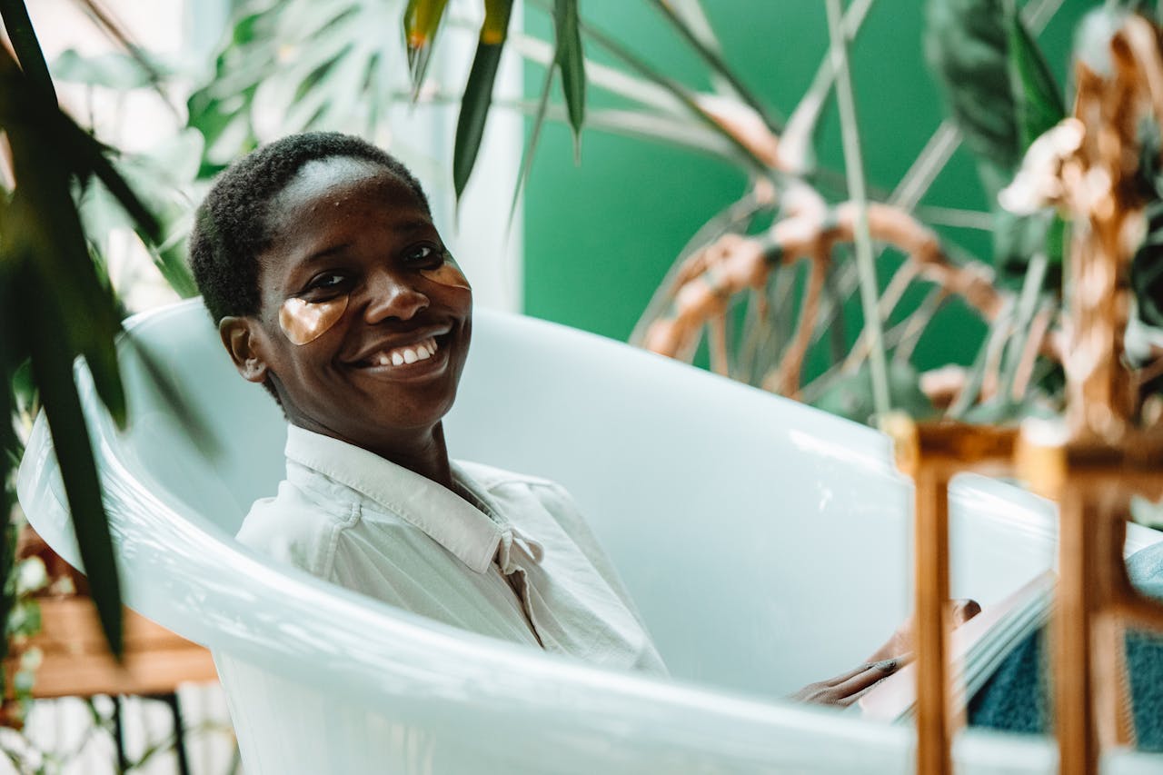 A joyful woman with under eye patches relaxes in a stylish bathtub amidst lush indoor plants.
